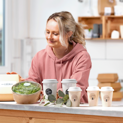 Una donna in un hoodie rosa sta preparando cibi sani, tra cui una ciotola di insalata e diverse tazze di caffè su un tavolo.