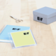 Ein blauer Notizblock, ein gelbes Notizblatt in einer transparenten Hülle, eine Schere und eine graue Geschenkbox auf einem Tisch.