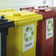 Drei Abfallbehälter für Recycling: gelb für Plastik, rot für Glas und schwarz für Restmüll, mit entsprechenden Symbolen.
