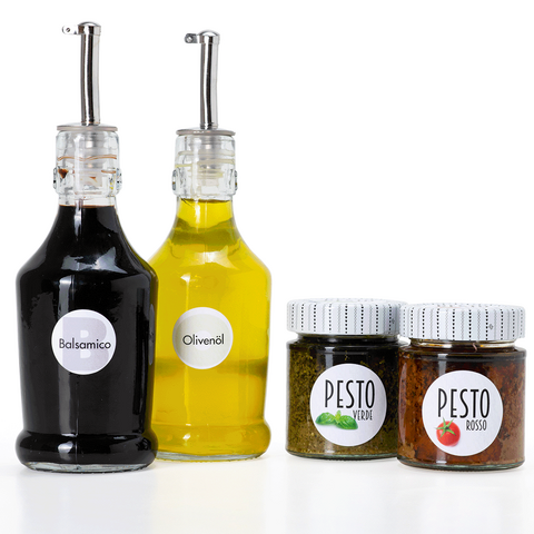 Due bottiglie di aceto balsamico e olio d'oliva, insieme a due barattoli di pesto in diverse varietà, su uno sfondo bianco.