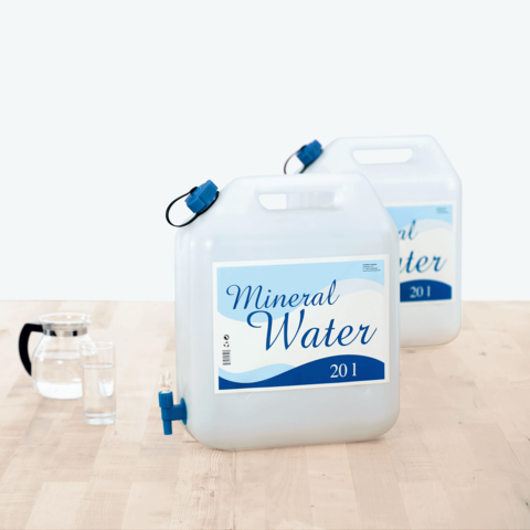 Zwei große Kanister mit Mineralwasser, jeweils 20 Liter, stehen auf einem Tisch neben einem Glas und einer Karaffe.