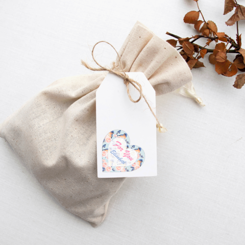Ein beige Stoffbeutel mit einem weißen Anhänger, der ein herzförmiges Etikett mit bunten Blumenmotiven zeigt.