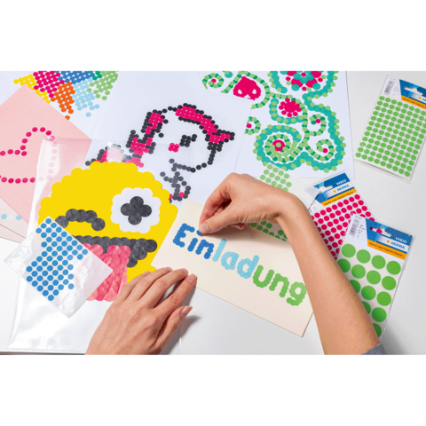 Eine Hand platziert bunte Punkte auf ein Einladungskarten-Design, um ein kreatives Kunstwerk zu gestalten. Verschiedene Bastelmaterialien sind sichtbar.