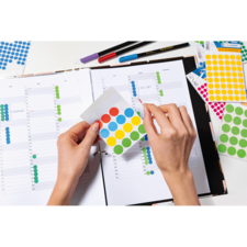 Eine Person hält ein Blatt mit bunten Aufklebern über einem geöffneten Planer, während sie eine Seite mit Kalenderwochen betrachtet.