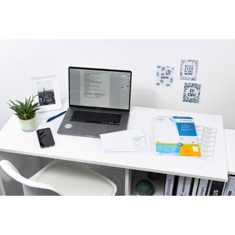 Schreibtisch mit einem Laptop, Smartphone, Pflanzen und Dokumenten. An der Wand hängen dekorative Notizen und Bilder.