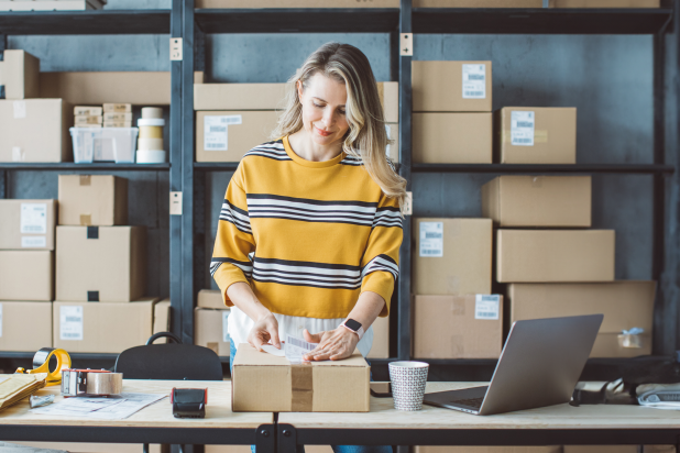 Una donna in un maglione a righe è in piedi a un tavolo, circondata da scatole e un laptop in un ufficio moderno.