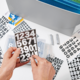 Hände halten eine transparente Box mit Zahlen und Buchstabenaufklebern, umgeben von weiteren Aufklebern und einem Ordner im Hintergrund.