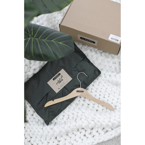 Ein schwarzes Paket mit einem Aufkleber, ein Holzkleiderbügel und eine weiße, gestrickte Decke. Eine Pflanze ist im Hintergrund sichtbar.