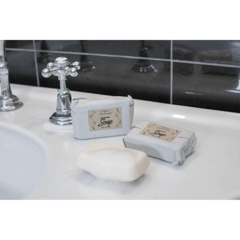 Zwei Seifenstücke, eines verpackt und eines unverpackt, liegen auf einer weißen Waschbeckenoberfläche neben einem Wasserhahn.