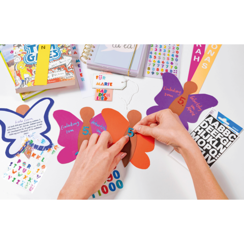 Hände gestalten bunte Papierfiguren mit Zahlen und Aufklebern auf einem Tisch, um Einladungen oder Dekorationen zu erstellen.