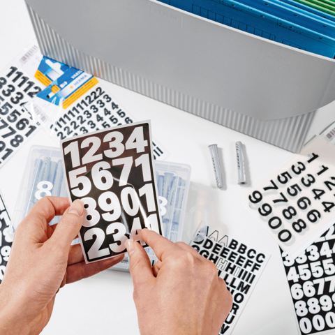 Hände halten eine transparente Box mit Zahlen und Buchstabenaufklebern, umgeben von verschiedenen Aufklebern auf einem Tisch.