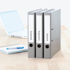 Drei graue Ordner mit Beschriftungen „Marketing“, „Controlling“ und „Rechnungen“ stehen auf einem Tisch neben einem Laptop und einem Notizblock.