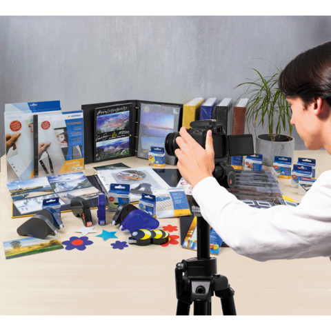 Eine Person fotografiert verschiedene Büroartikel und Präsentationsmaterialien, die auf einem Tisch angeordnet sind. Im Hintergrund sind einige Ordner und eine Pflanze sichtbar.