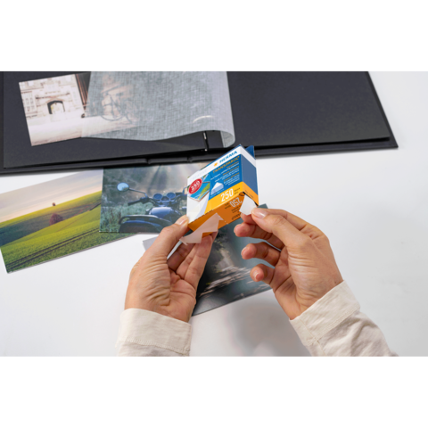 Eine Person hält eine Verpackung mit Fotopapier in den Händen, während mehrere Fotos und ein Album auf einem Tisch liegen.