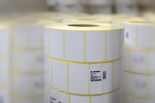 Rulli con etichette bianche, impilati in un ambiente di magazzino. Le etichette sono contrassegnate con linee gialle.