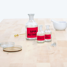 Flaschen mit roten Etiketten, beschriftet mit "Spir. Aether", "Barbital" und "Tinct. Tramon", sowie ein Mörser und zwei Glasplatten auf einem Tisch.