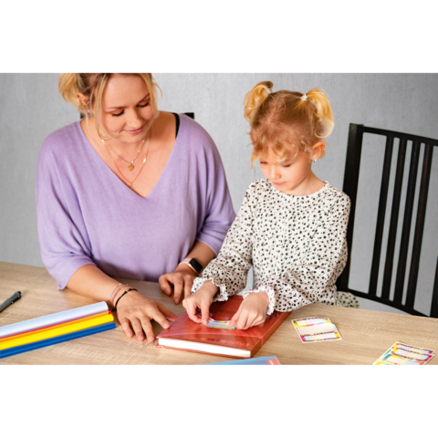 Eine Frau und ein kleines Mädchen sitzen an einem Tisch, während sie gemeinsam an einem Buch arbeiten. Bunte Stifte und Karten liegen daneben.