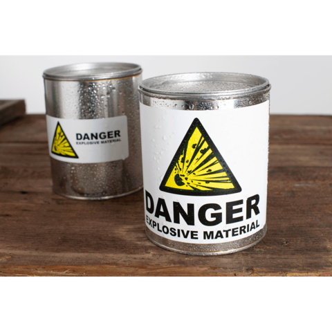 Metallbehälter mit Warnaufkleber „Gefahr explosive Materialien“ auf einem Holztisch. Ein weiterer Behälter im Hintergrund.