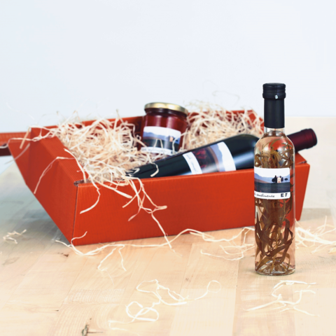 Ein roter Geschenkkorb mit einer Flasche Likör, einem Glas Marmelade und einer Flasche Wein, umgeben von Holzwolle.