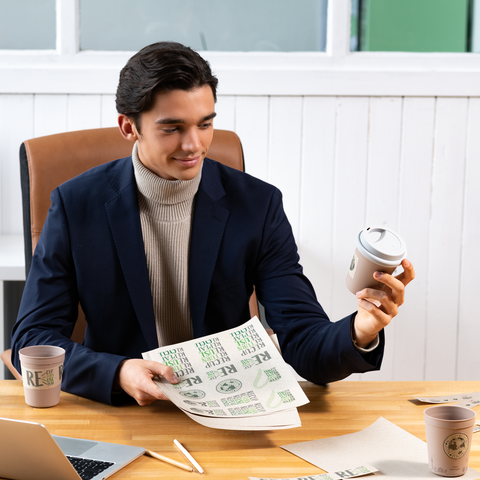 Ein junger Mann in einem Anzug hält einen Kaffeebecher und betrachtet Papiere auf einem Tisch. Zwei Kaffeetassen stehen ebenfalls auf dem Tisch.
