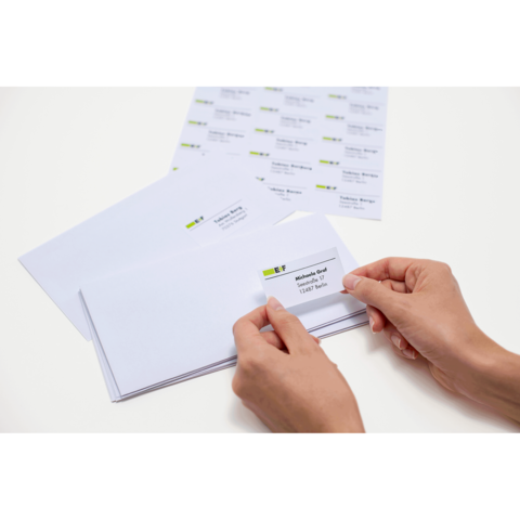 Eine Person klebt einen Adressaufkleber auf einen Umschlag, während mehrere leere Umschläge und ein Blatt mit Aufklebern auf dem Tisch liegen.