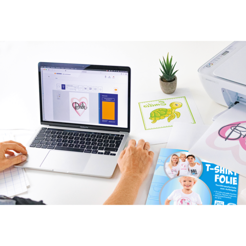 Eine Person arbeitet an einem Laptop, um ein Design für T-Shirt-Folie zu erstellen. Neben dem Laptop liegen Druckmaterialien und eine kleine Pflanze.