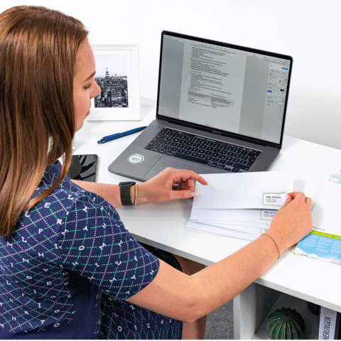 Eine Person sitzt an einem Schreibtisch und arbeitet am Laptop, während sie einen Brief in der Hand hält. Dokumente und Büromaterial sind sichtbar.