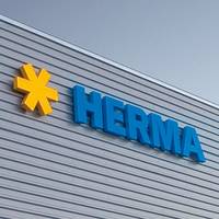 Gelbe Stern-Logos und blaue Buchstaben bilden das Wort "HERMA" an einer modernen Gebäudeaußenwand.