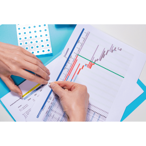 Eine Hand platziert einen Aufkleber auf ein Diagramm, das Daten und Statistiken zeigt, während ein Blatt Papier und ein farbiger Hintergrund sichtbar sind.