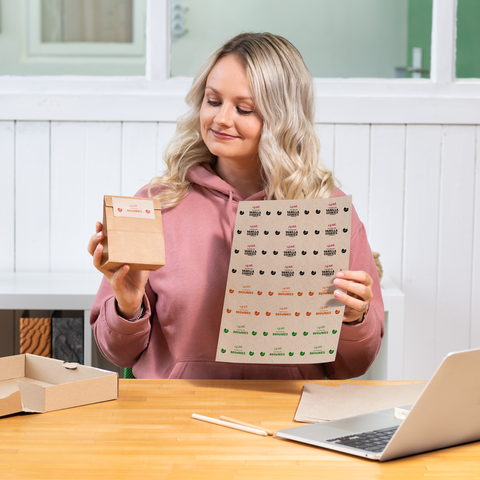 Eine Person mit blonden Haaren hält eine kleine Verpackung und ein Blatt mit Aufklebern in einer hellen Umgebung.