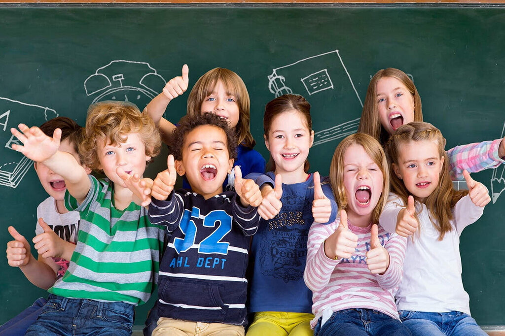 Un gruppo di bambini è davanti a una lavagna, sorride e posa felicemente. Disegni di materiale scolastico sono visibili sullo sfondo.