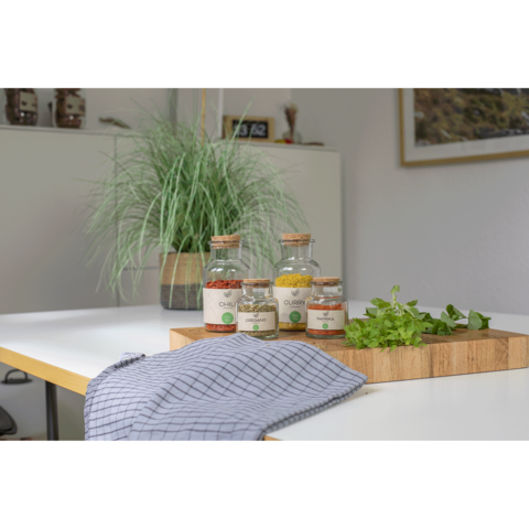 Gläser mit Gewürzen und frischen Kräutern auf einem Holzbrett, daneben ein kariertes Geschirrtuch und eine grüne Pflanze im Hintergrund.