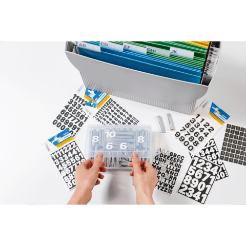 Hände halten eine transparente Box mit Zahlen und Buchstabenaufklebern, umgeben von weiteren Aufklebern und einem Ordner mit farbigen Trennblättern.