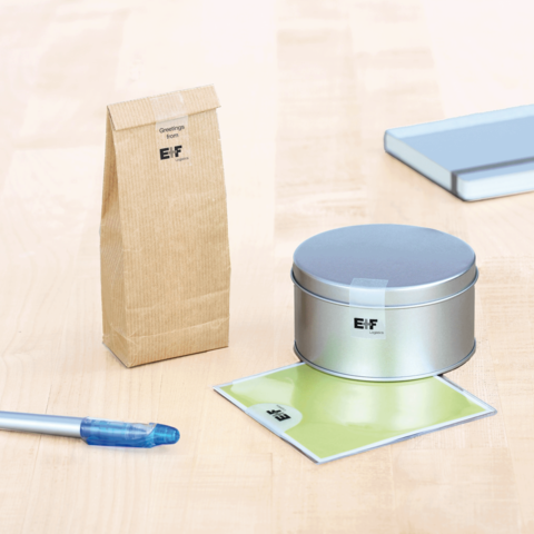 Ein brauner Papierbeutel, eine silberne Dose, ein grünes Blatt und ein blauer Stift auf einem Tisch. Alle Objekte sind mit dem Logo "E.F." versehen.