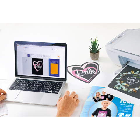 Eine Hand bedient einen Laptop, auf dem ein Design mit einem Herz und dem Wort "Bride" angezeigt wird. Neben dem Laptop steht ein Drucker.