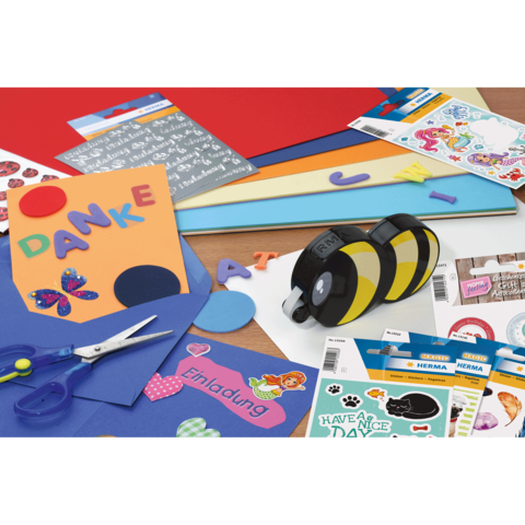 Bunte Bastelmaterialien auf einem Tisch, darunter Papier, Aufkleber, Scheren und ein doppelseitiger Kleberoller.
