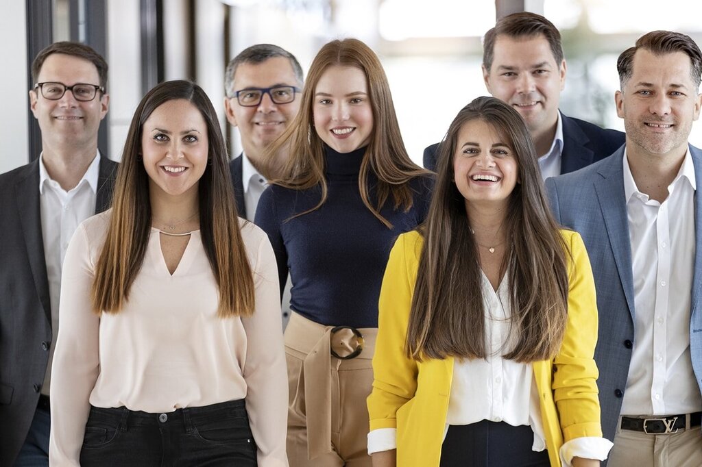 Un gruppo di sette persone si trova in un corridoio, sorridendo e vestite in modo formale; alcune indossano camicie e completi.