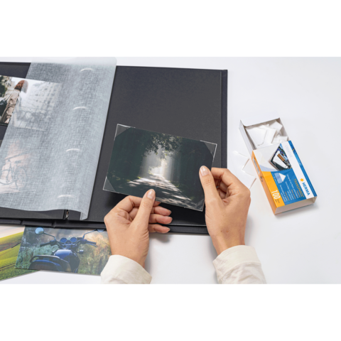 Eine Person hält ein Foto und fügt es in ein Fotoalbum ein, während sich daneben eine Verpackung mit Fotoecken befindet.
