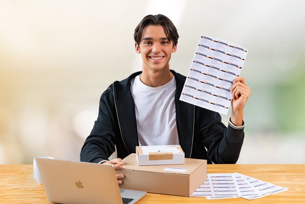 Un giovane uomo è seduto a un tavolo con un laptop e tiene in mano un foglio con adesivi.