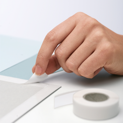 Una mano rimuove un pezzo di nastro adesivo da una superficie grigia, mentre un altro pezzo di nastro adesivo giace accanto.