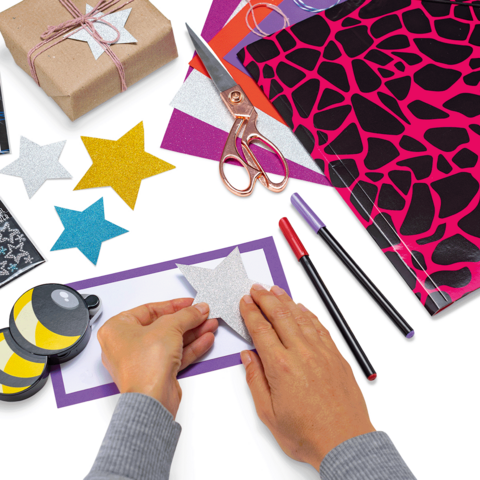 Mani che incollano una stella luccicante su un foglio colorato, circondate da materiali per il bricolage come forbici, pennarelli e carta da regalo.