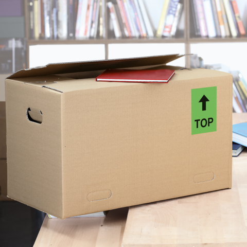 Ein Karton mit dem Aufkleber "FRAGILE", daneben ein graues Kuvert mit "DRINGEND!" und eine weiße Karte. Schere liegt ebenfalls auf dem Tisch.