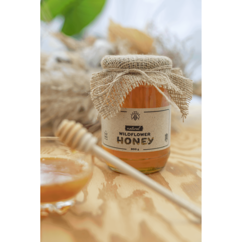 Glas Honig mit Jute-Deckel und Etikett "Wildflower Honey" neben einem Honiglöffel und einer kleinen Schale mit Honig.