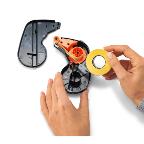 Eine Hand hält ein Stück gelbes Klebeband neben einem abgenommenen Gehäuse eines Klebebandabrollers mit sichtbaren Zahnrädern.
