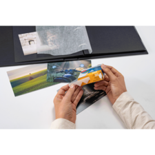 Eine Person hält eine Packung Fotohüllen und betrachtet mehrere Fotos auf einem Tisch, während ein Fotoalbum im Hintergrund sichtbar ist.