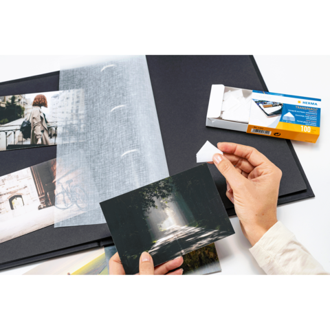 Eine Person hält eine Fotografie in der Hand, während sie ein Fotoalbum mit weiteren Bildern und einer Packung Fotohüllen betrachtet.