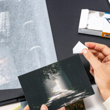 Eine Person hält eine Fotografie in der Hand, während sie ein Fotoalbum mit weiteren Bildern und einer Packung Fotohüllen betrachtet.